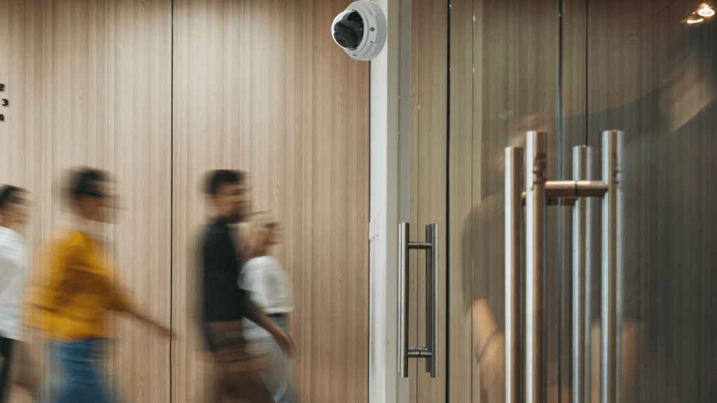 Entryway with glass doors and people blurry walking by, with an Axis camera by the doorway.