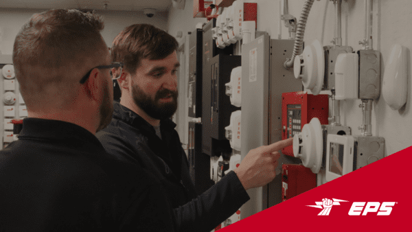 Two men discussing on the left hand side, one man pointing to the right side of the image where there is a smoke detector surrounded by a wall of electrical boxes and security test equiptment. There are CO detectors, a large fire panel, smart home systems with touchscreens, motion sensors, and larger commercial panels in the background for demo equipment.